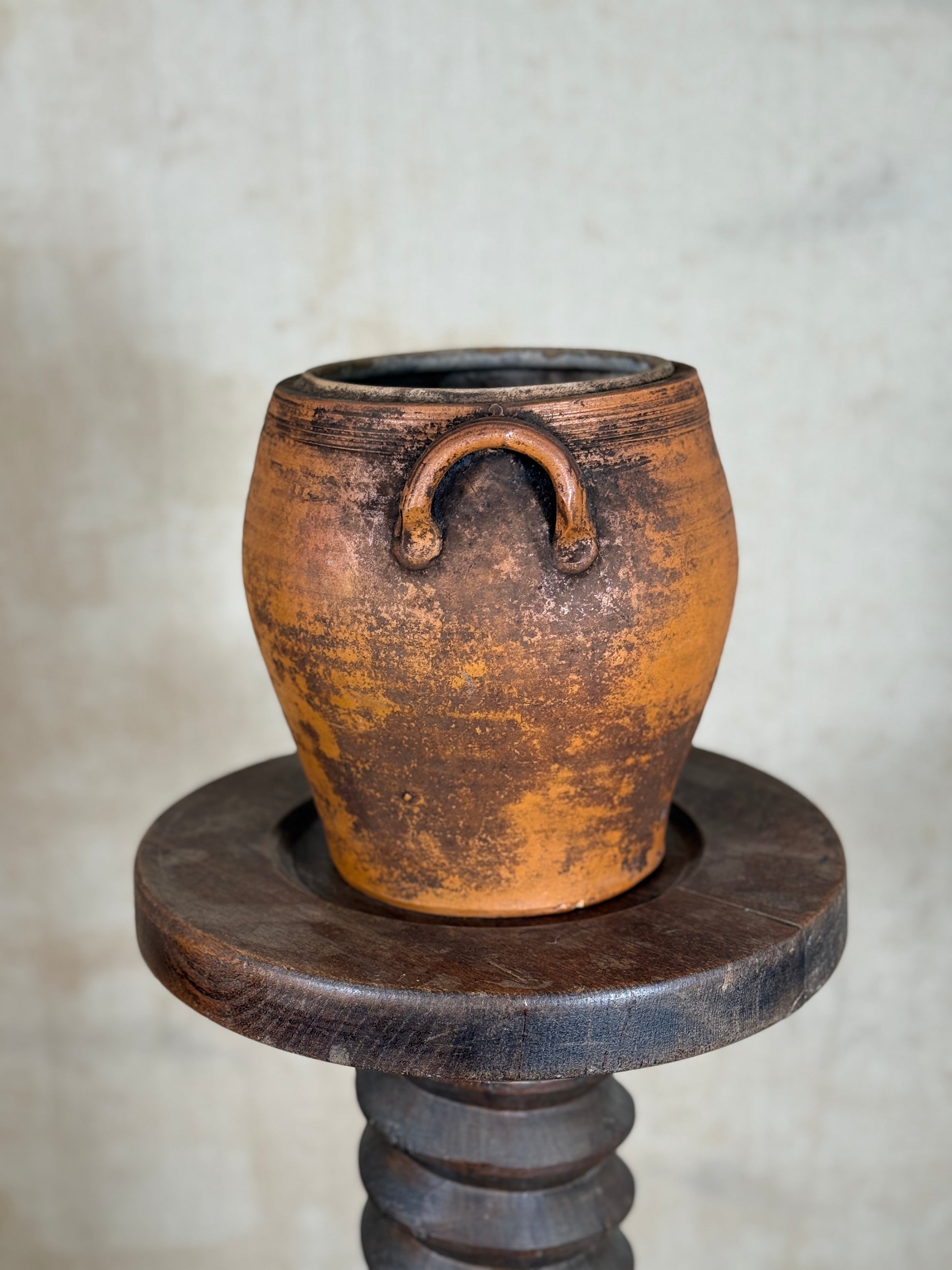 A rustic terracotta clay pot with a single loop handle, displayed on a dark wooden pedestal against a neutral textured wall.