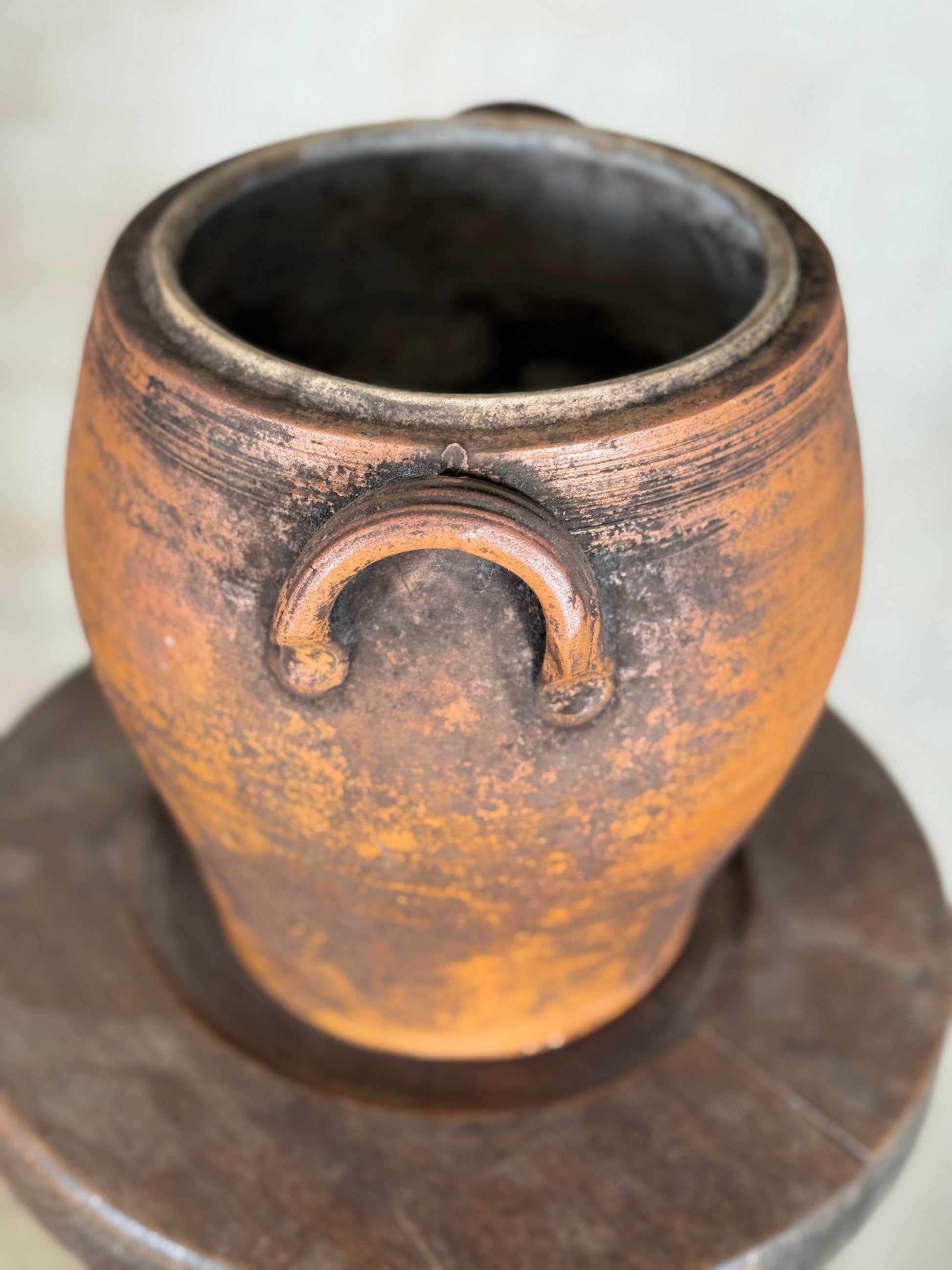A rustic terracotta clay pot with two loop handles, displayed on a dark wooden stand against a light beige background.