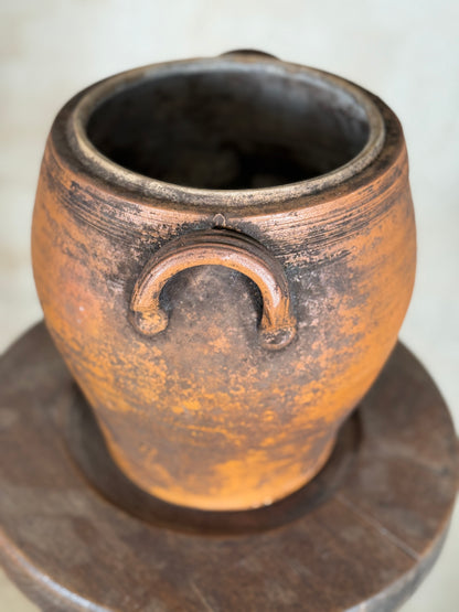 A rustic terracotta clay pot with two loop handles, displayed on a dark wooden stand against a light beige background.