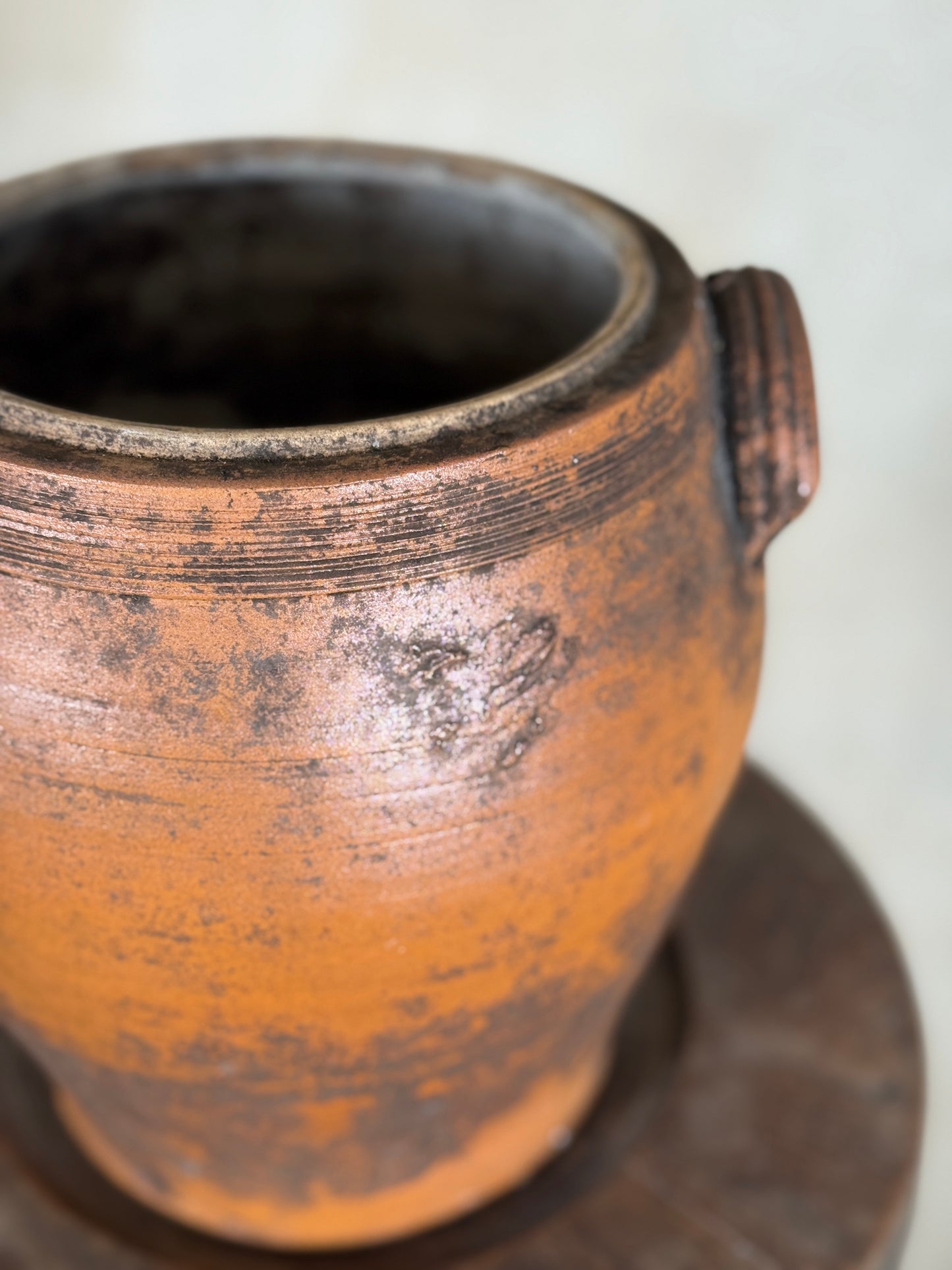 A close-up of a rustic terracotta clay pot with a dark brown handle and weathered, distressed finish, sitting on a dark wooden surface against a light beige background.