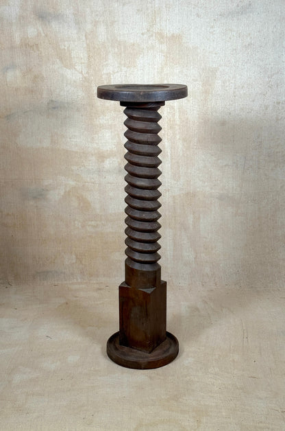 A rustic industrial stool or pedestal made from a large threaded screw bolt, featuring a round wooden seat on top, a threaded brown metal stem, a hexagonal base, and a circular foot ring, set against a textured beige backdrop.