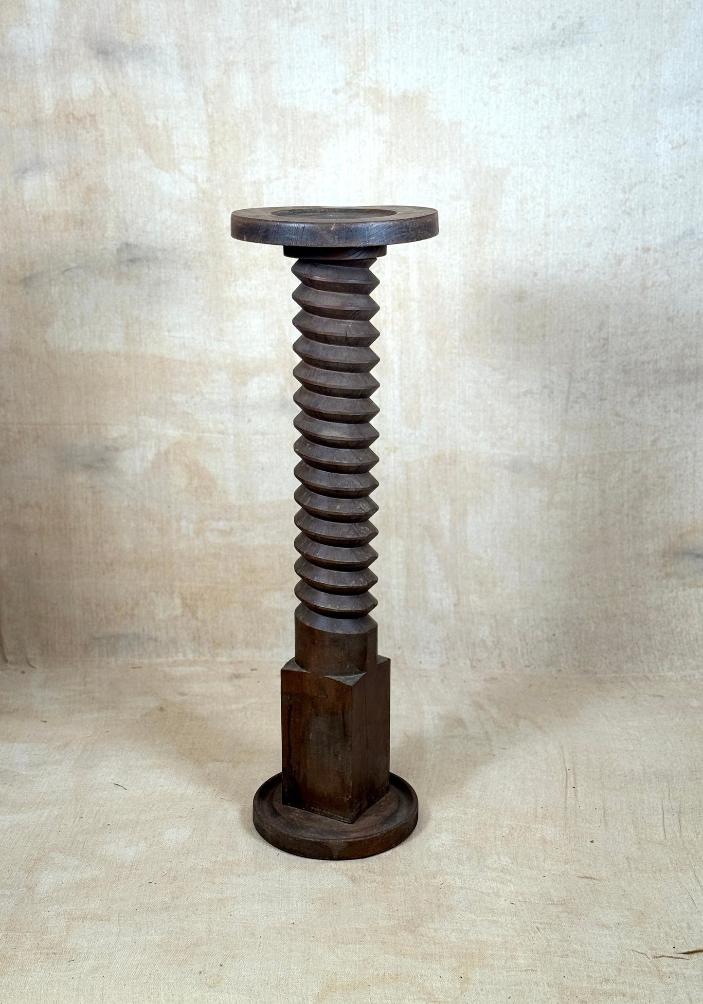 A rustic dark wood pedestal stand shaped like a giant threaded screw, featuring a circular top and hexagonal base, set against a textured beige backdrop.