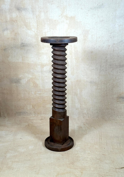 A rustic dark wood pedestal stand shaped like a giant threaded screw, featuring a circular top and hexagonal base, set against a textured beige backdrop.