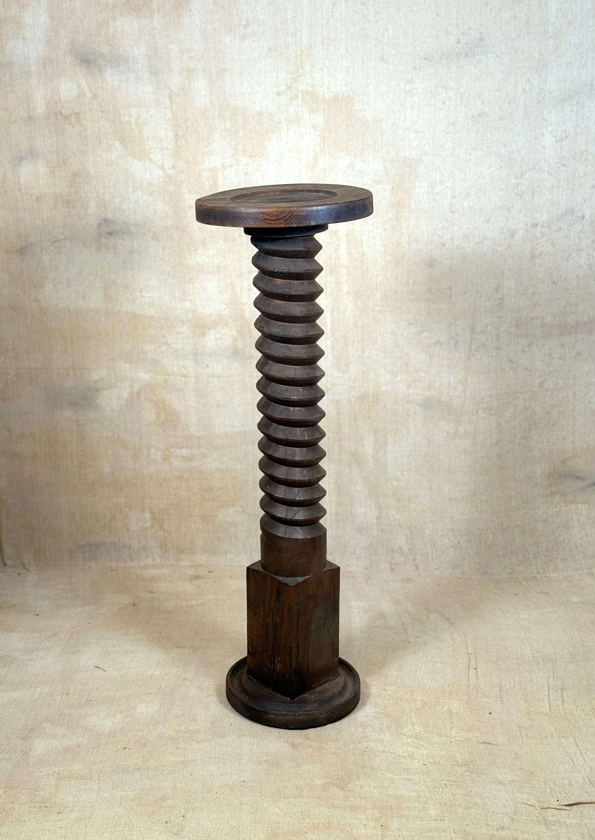 A dark brown wooden sculpture or stand shaped like a giant threaded screw, standing upright against a textured beige backdrop.