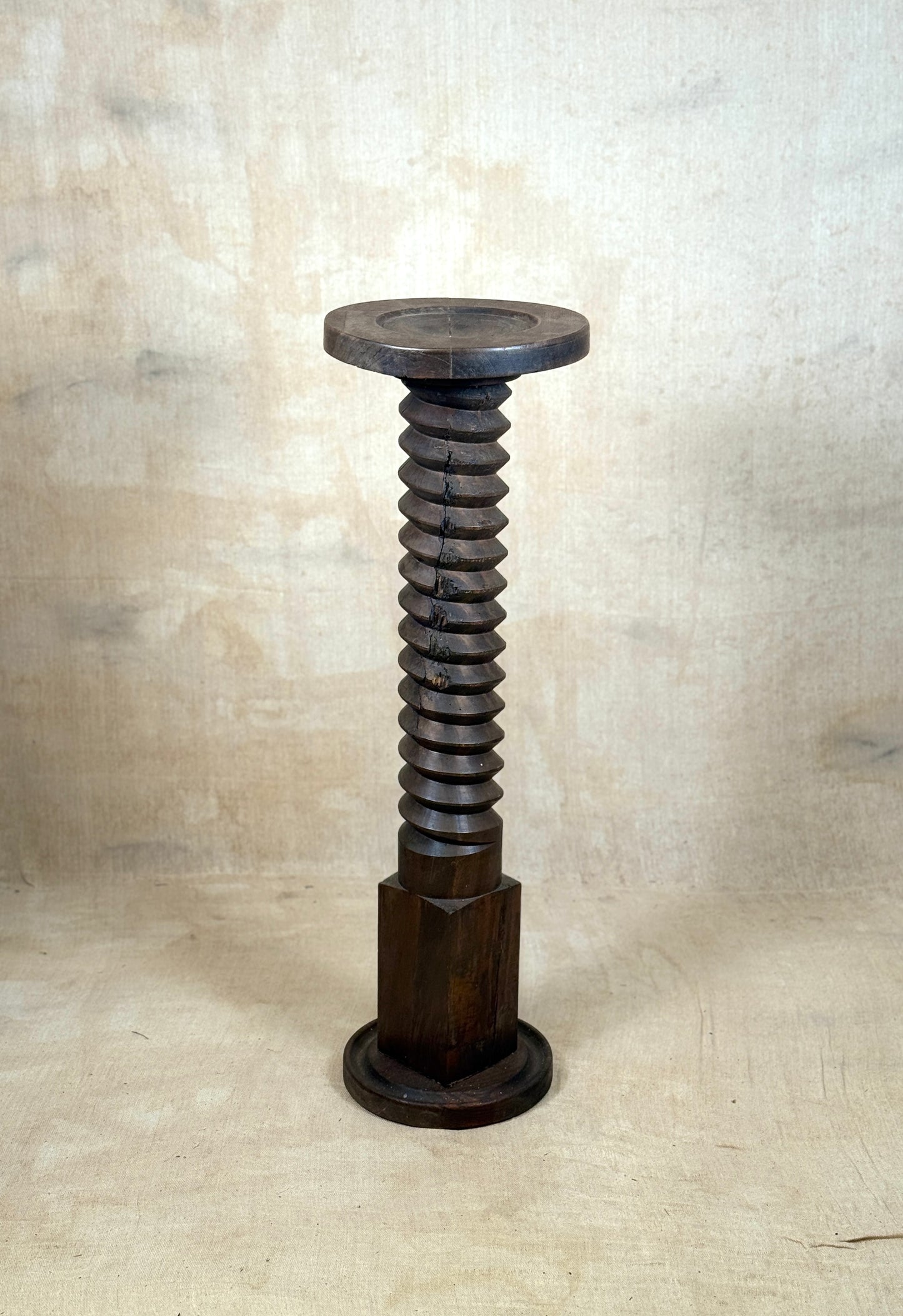 A dark brown wooden sculpture shaped like a giant threaded screw bolt with a circular top and base, set against a textured beige backdrop.