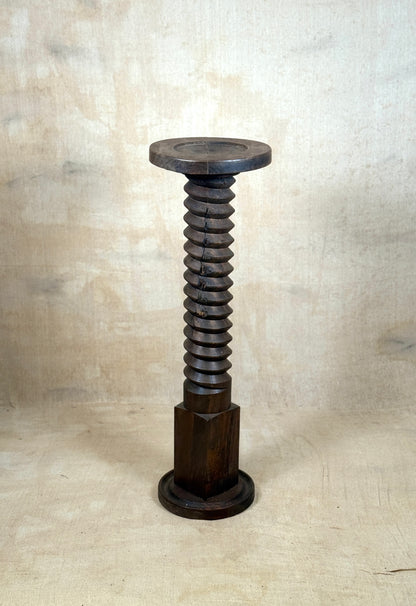 A dark brown wooden sculpture shaped like a giant threaded screw bolt with a circular top and base, set against a textured beige backdrop.