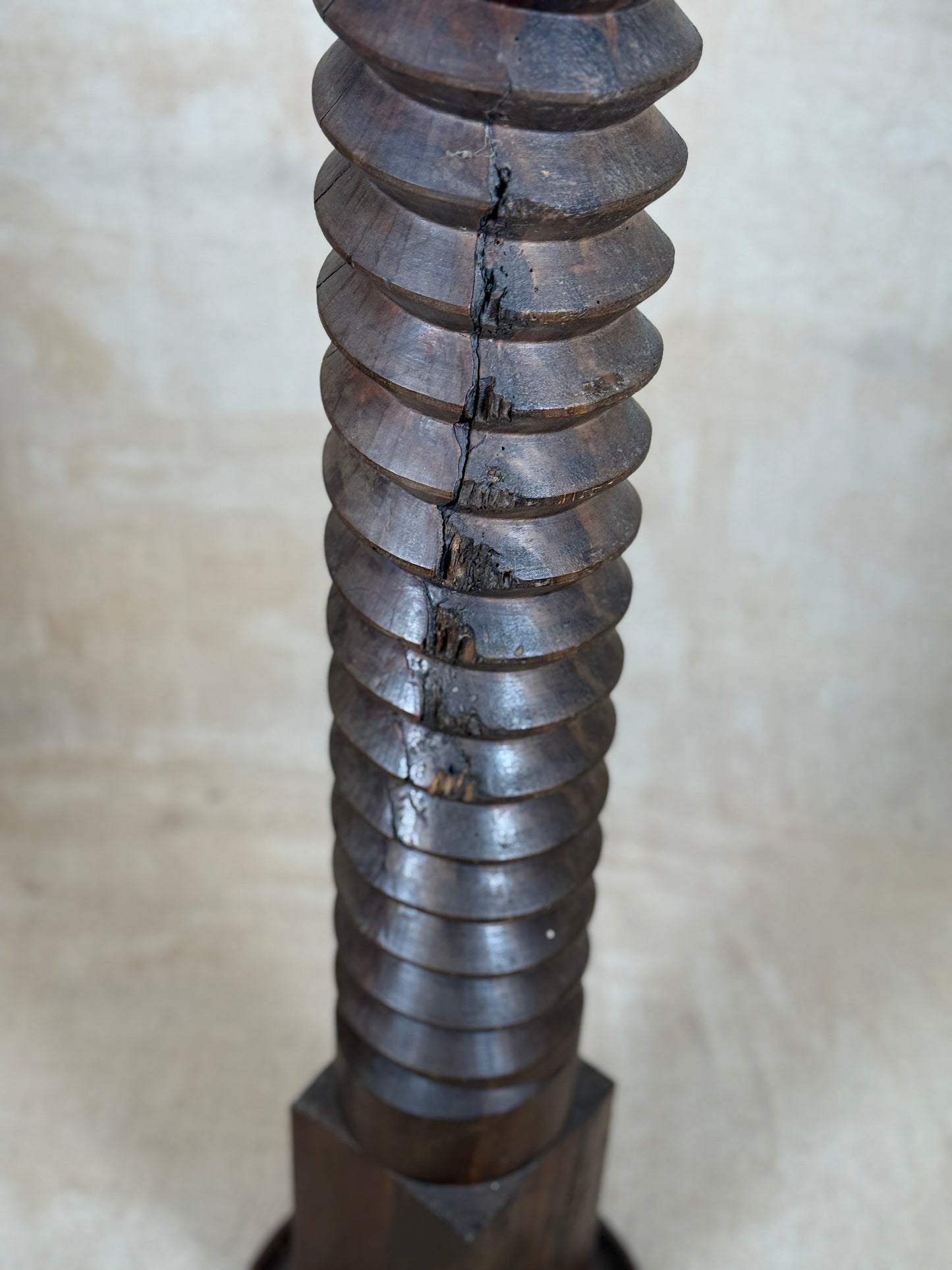 A close-up of a dark brown wooden screw or threaded post with a visible crack running vertically down the center, set against a neutral beige background.