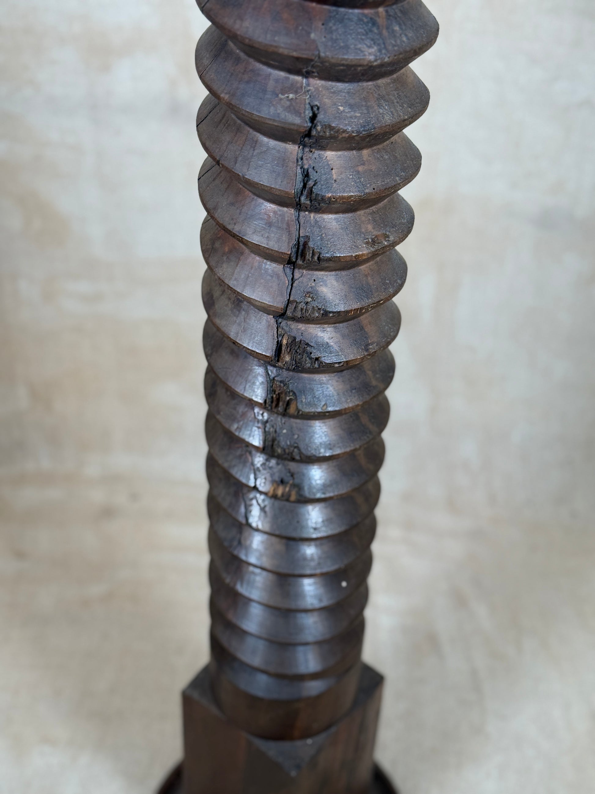 A close-up of a dark brown wooden screw or threaded post with a visible crack running vertically down the center, set against a neutral beige background.