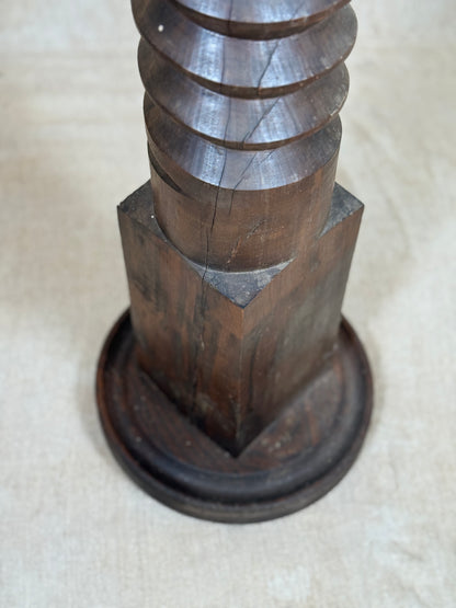 A close-up of a dark brown wooden decorative object with a fluted cylindrical top and a round pedestal base, photographed on a light beige fabric surface.