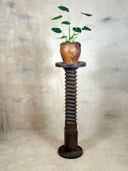 A rustic dark brown wooden pedestal stand shaped like a threaded screw, holding a terracotta pot with green plant leaves, set against a textured beige fabric backdrop.
