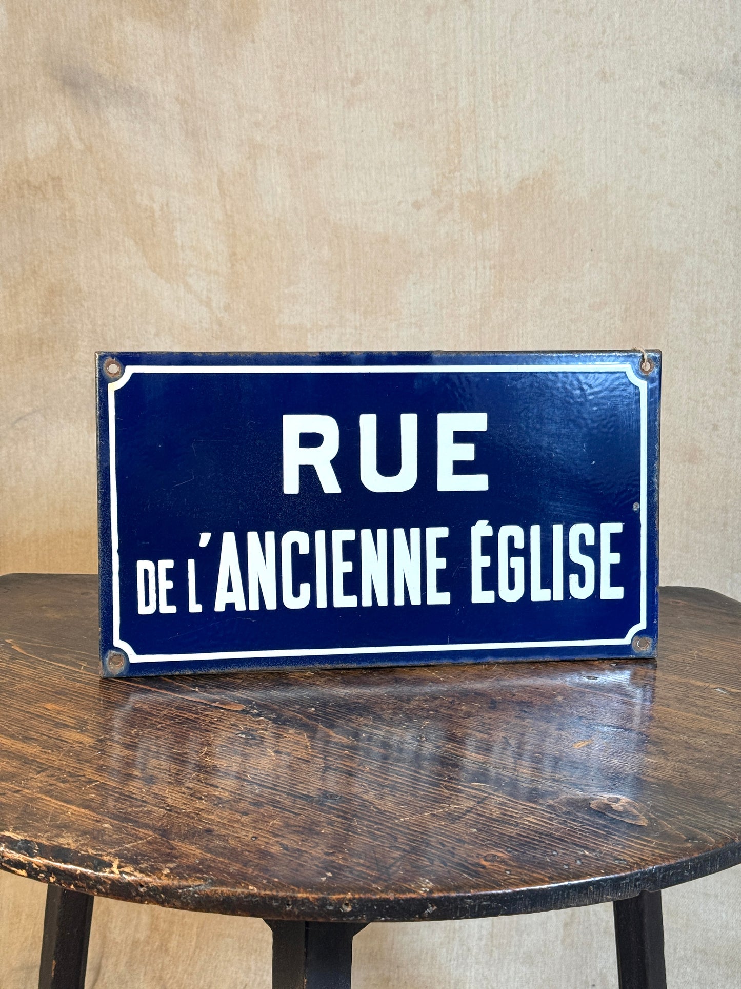 A rectangular vintage-style blue street sign with white French text reading 'RUE DE L'ANCIENTE EGLISE', displayed on a dark wooden table against a beige textured wall.
