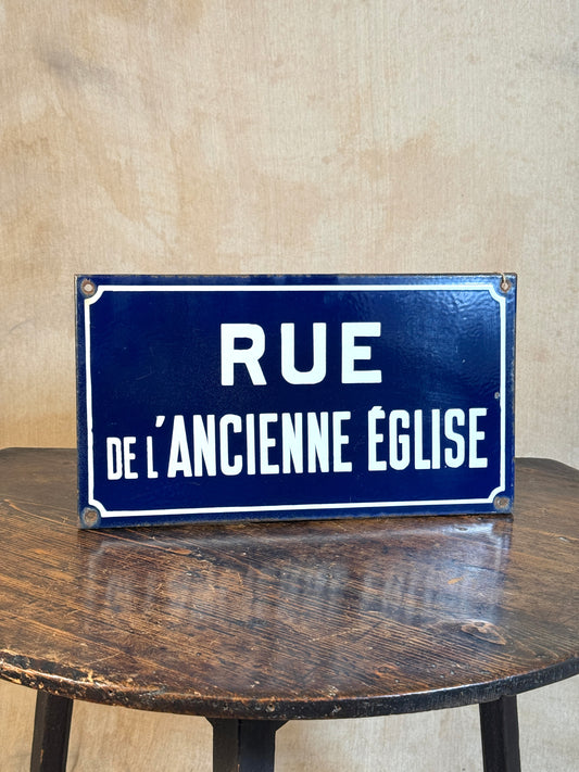 A rectangular vintage-style blue street sign with white French text reading 'RUE DE L'ANCIENTE EGLISE', displayed on a dark wooden table against a beige textured wall.