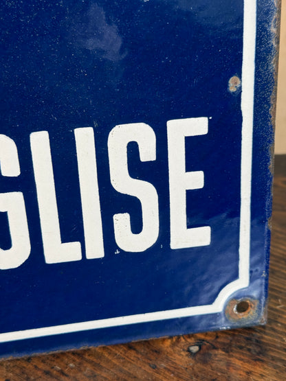 A close-up of a vintage blue enamel sign with white lettering and a white border, showing part of the word 'ENGLESE' on a rustic wooden surface.
