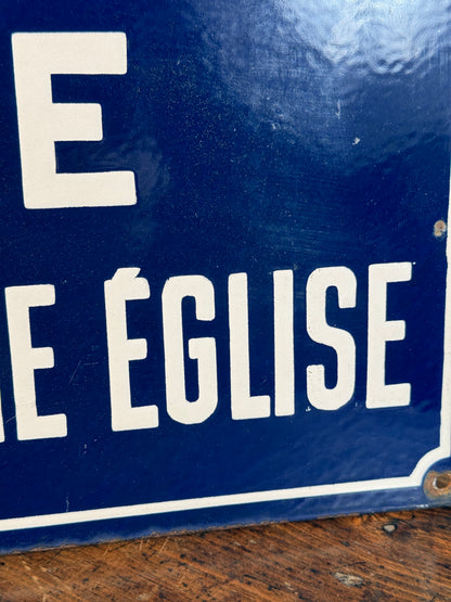 A close-up of a blue enamel sign with bold white French text reading 'EGLISE' vertically, showing some wear and a wooden surface in the background.