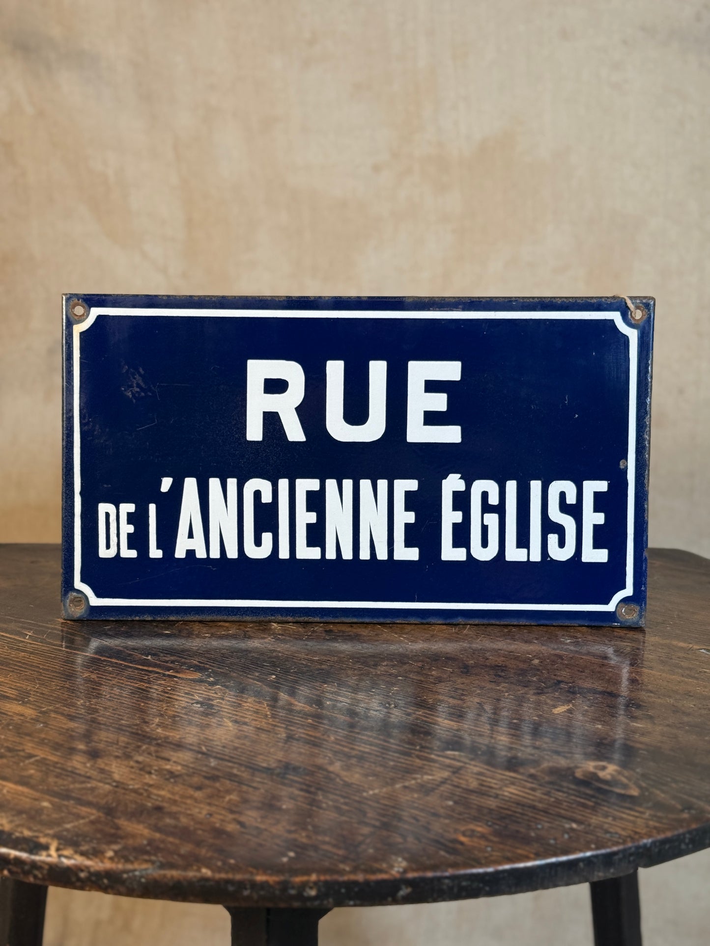 A vintage-style rectangular blue sign with white lettering reading 'RUE DE L'ANCIENTE EGLISE', displayed on a dark wooden table against a textured beige wall.