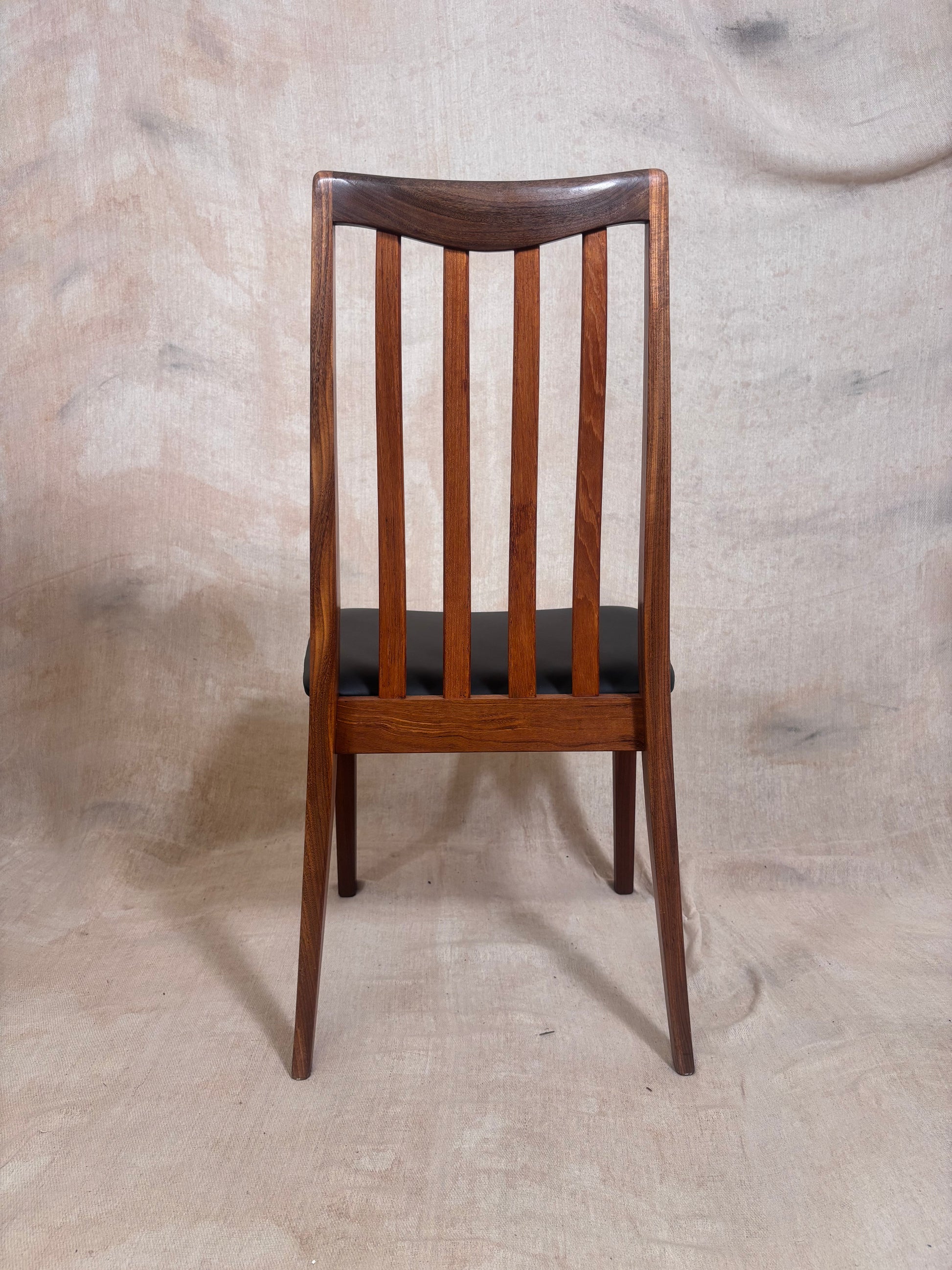 A mid-century modern wooden dining chair with a warm brown wood frame, vertical slats, and a black upholstered seat, shown against a neutral textured beige backdrop.