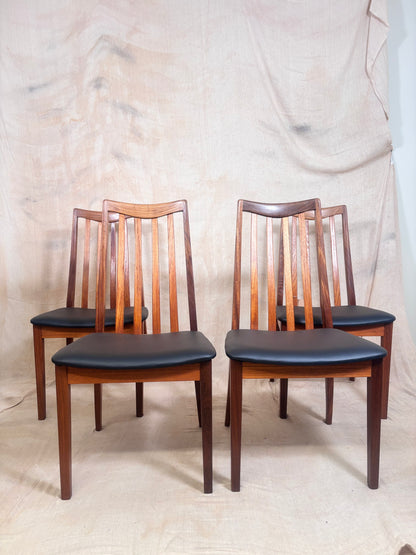 Four wooden dining chairs with black upholstered seats, arranged in two pairs against a draped beige fabric backdrop.