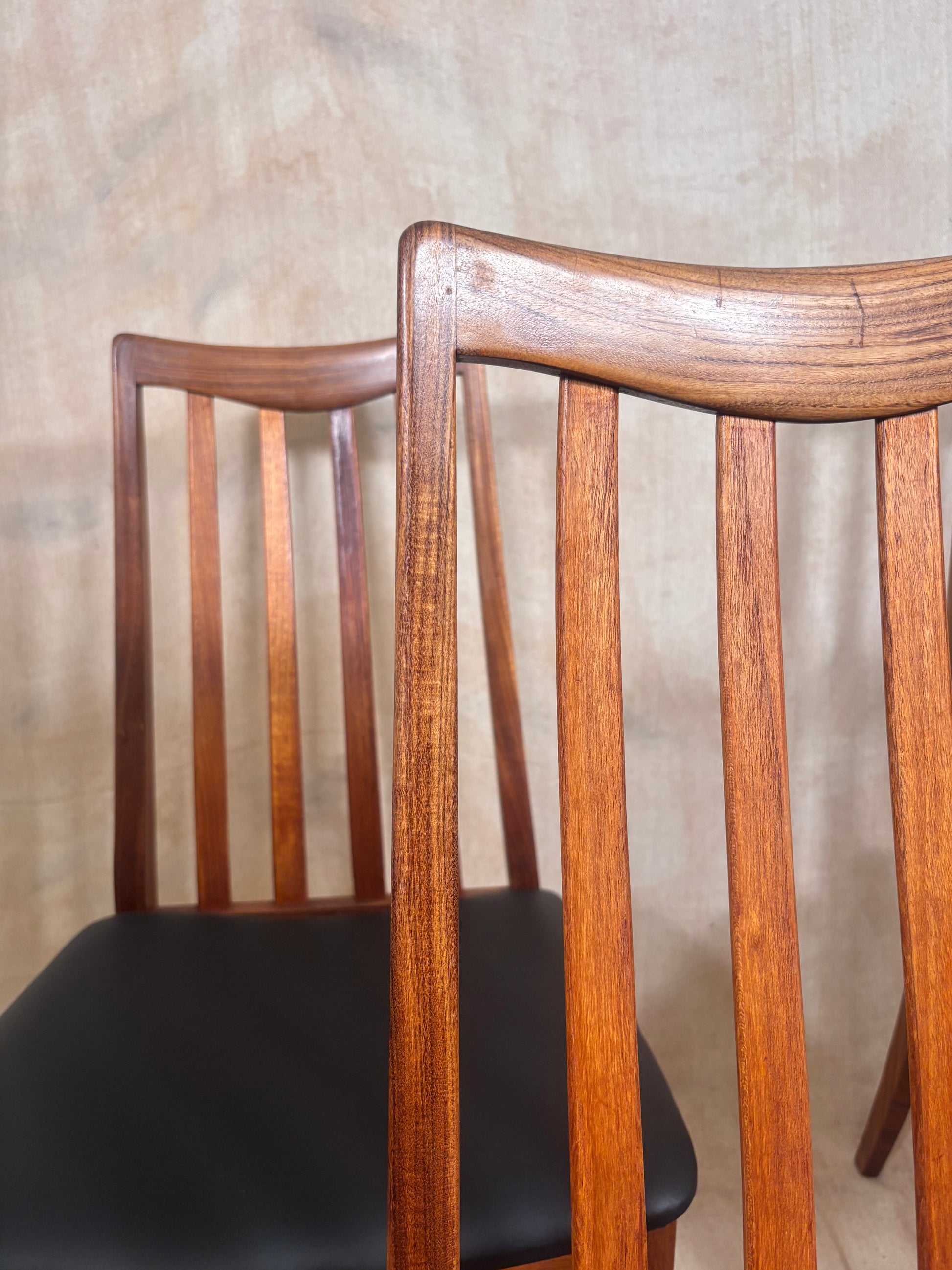A close-up of a mid-century modern wooden dining chair with a warm brown wood frame and a black upholstered seat, set against a textured beige wall.