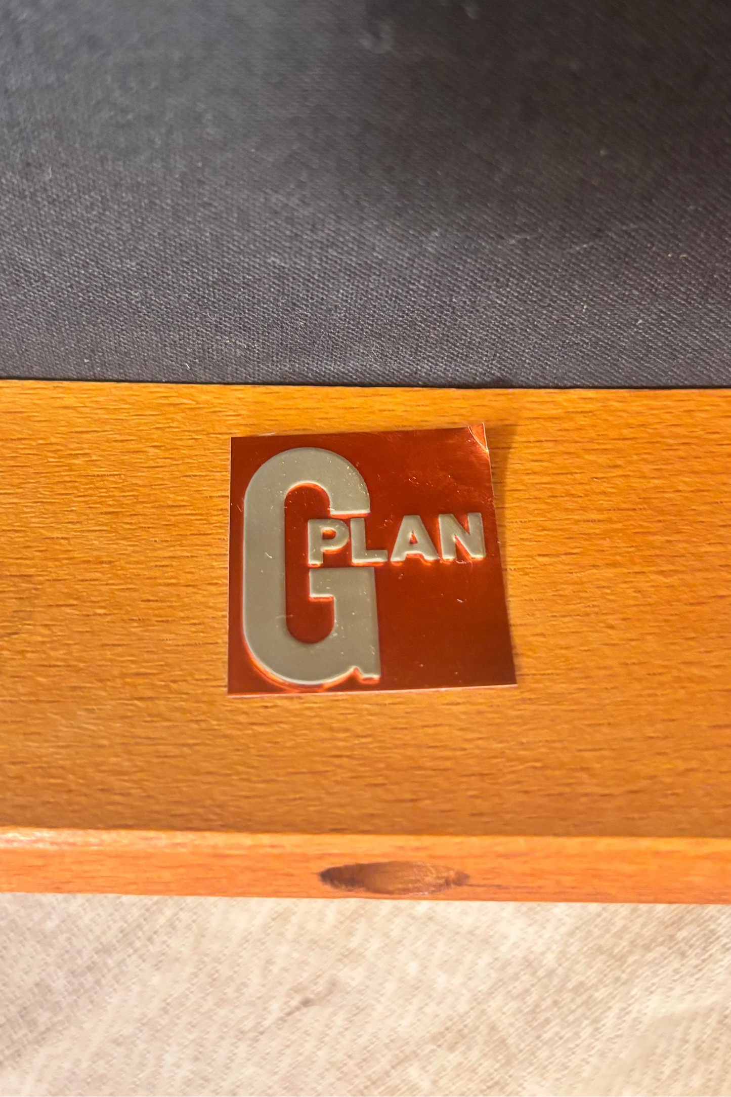 Close-up of a wooden surface with a small metallic red and silver 'G PLAN' emblem, next to a dark gray textured panel.