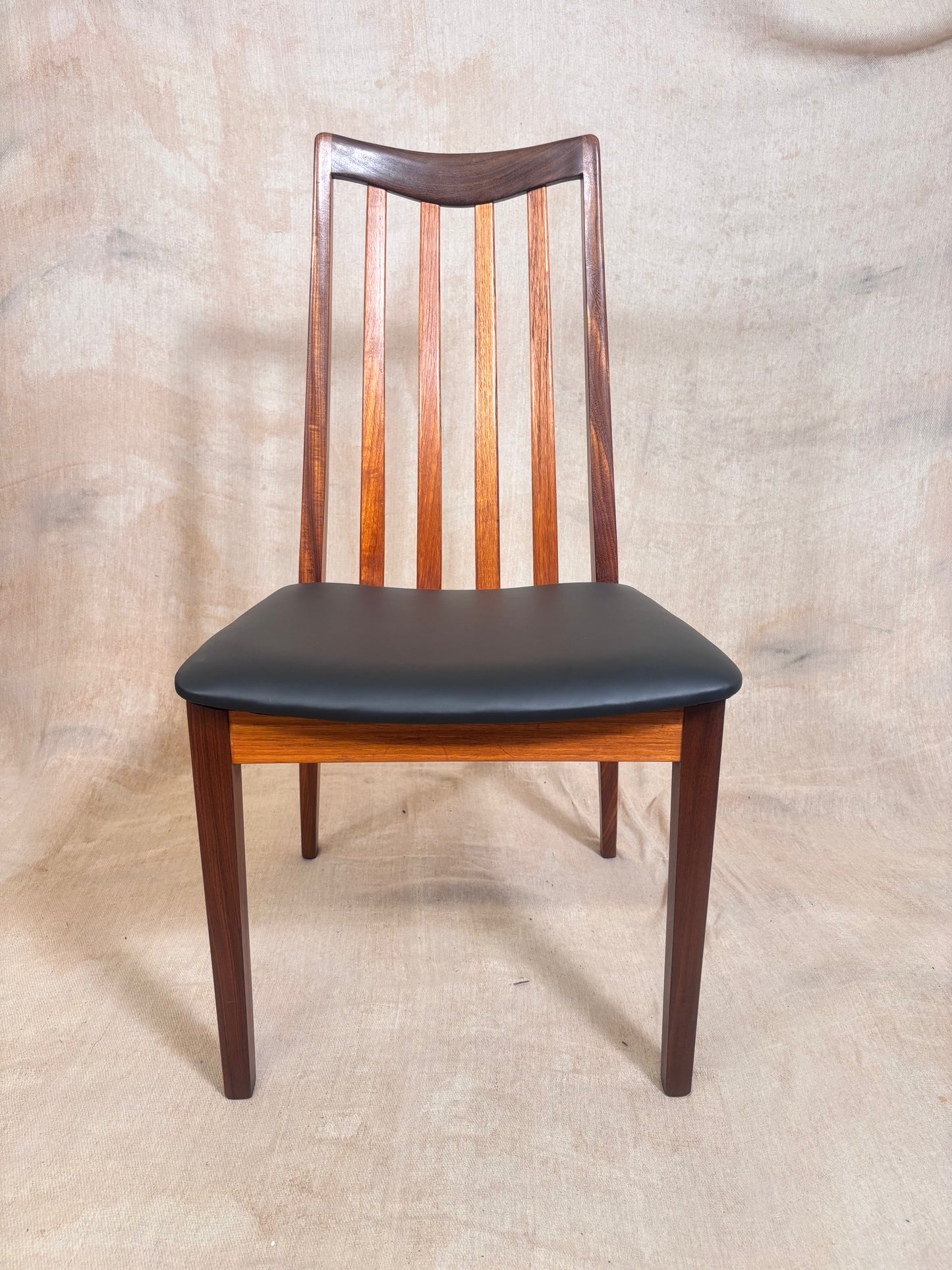 A mid-century modern wooden dining chair with a curved backrest, vertical slats, a warm medium-brown wood frame, and a black upholstered seat cushion, set against a neutral textured beige backdrop.