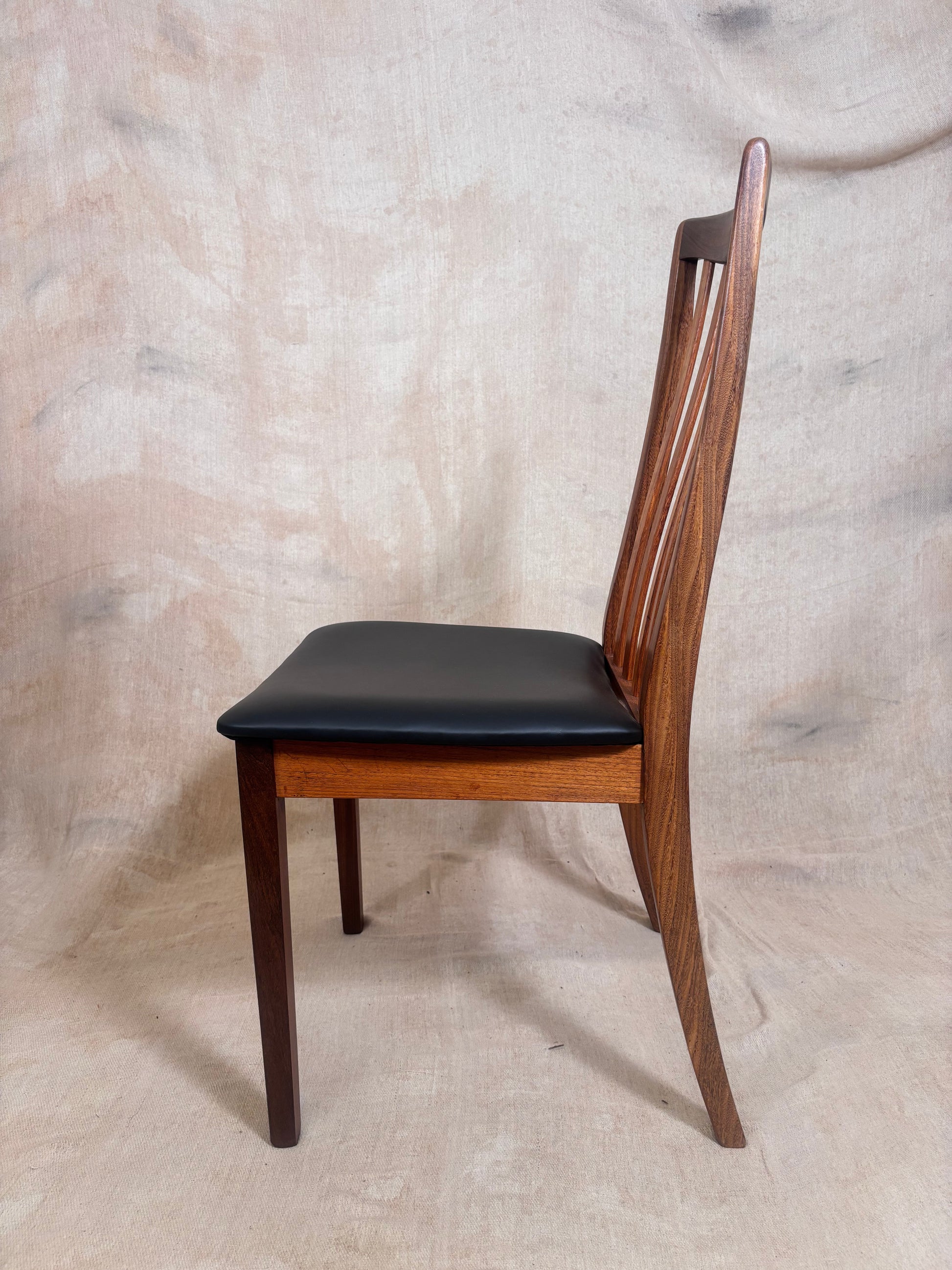A mid-century modern wooden dining chair with a warm brown wood frame and a black upholstered seat, shown from a side profile against a textured beige fabric backdrop.