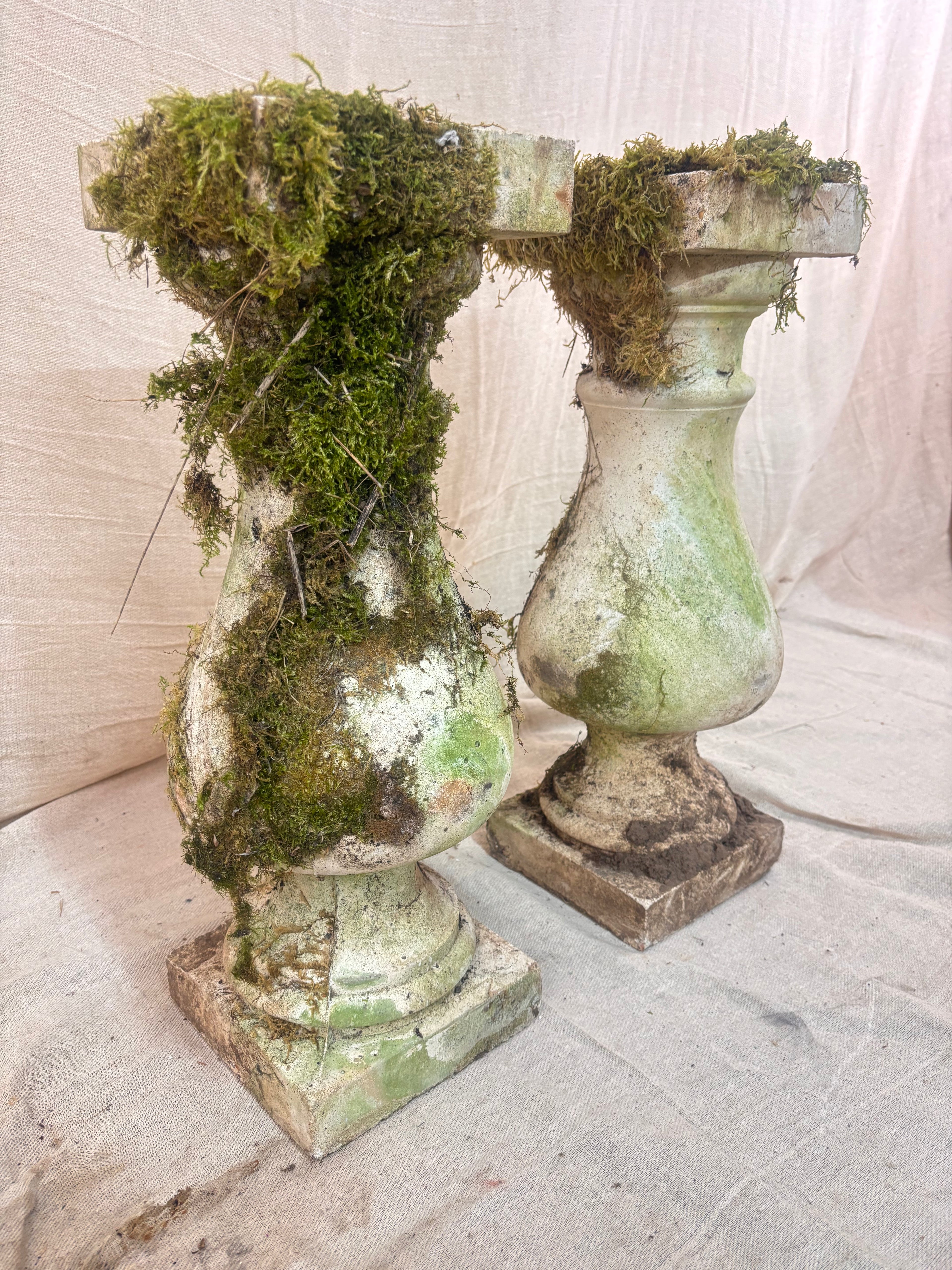 Vintage stone garden pedestals with a classic baluster shape, stepped square bases, and flat square tops, finished in weathered pale gray stone with natural moss and lichen growth.