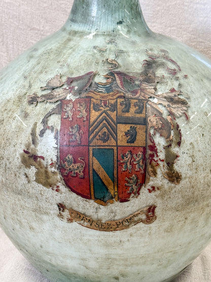 Close-up of an antique glass demijohn bottle with a rounded body, a narrow neck, and a hand-painted heraldic coat of arms on a distressed, aged off-white glaze.