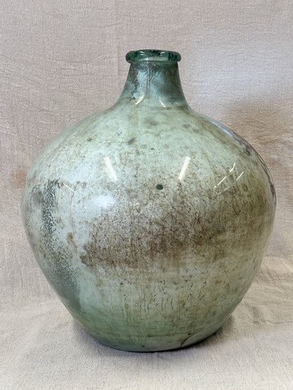 A large victorian glass carboy with a bulbous rounded body and a short neck, featuring a distressed glaze in muted green, beige, and gray tones, set against a neutral fabric backdrop.