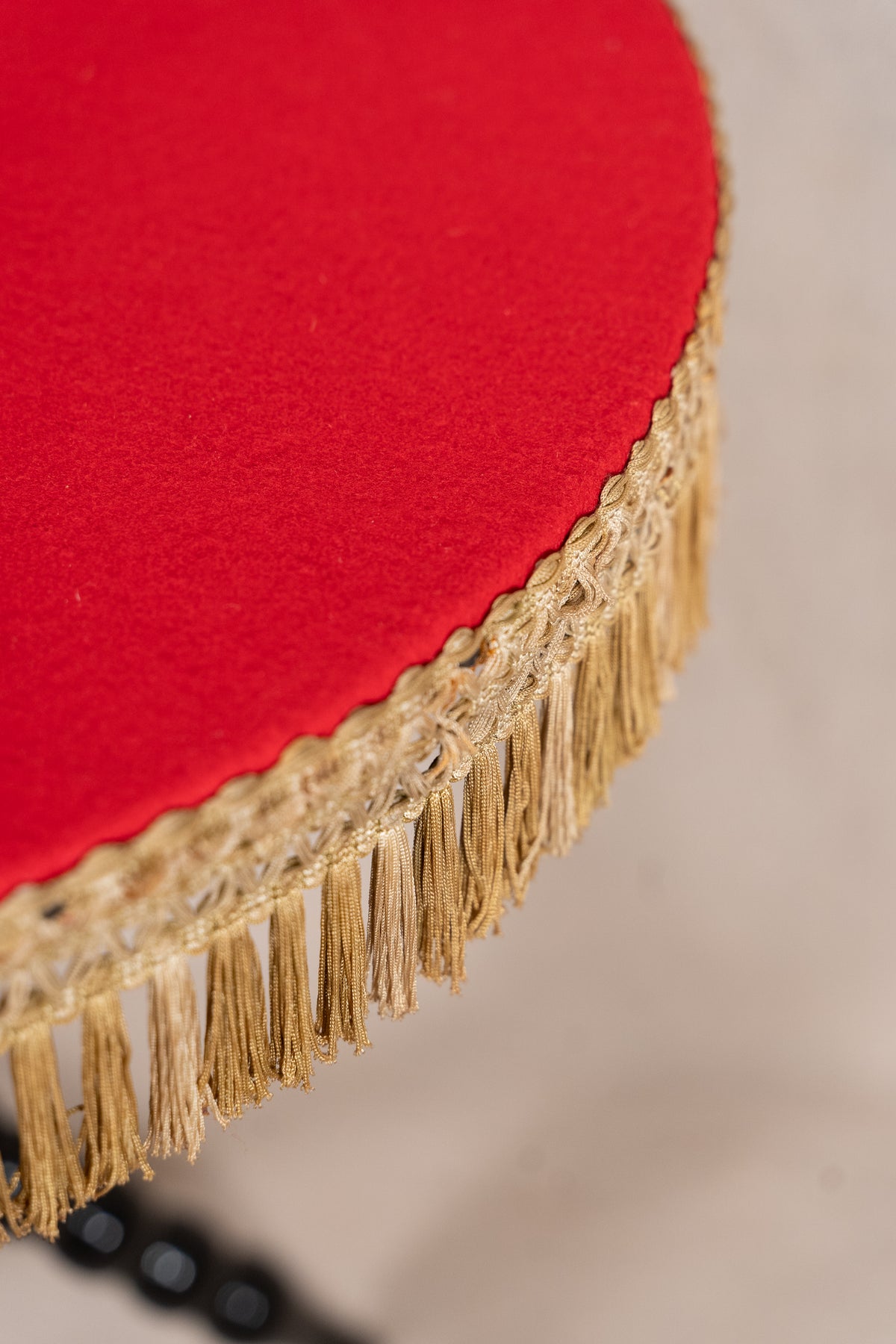 Close-up of a round red upholstered stool or chair with a decorative beige fringed trim around the edge, set against a light neutral background.