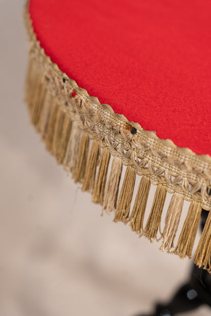 Close-up of a round red upholstered stool or chair with ornate gold fringe trim around the edge.