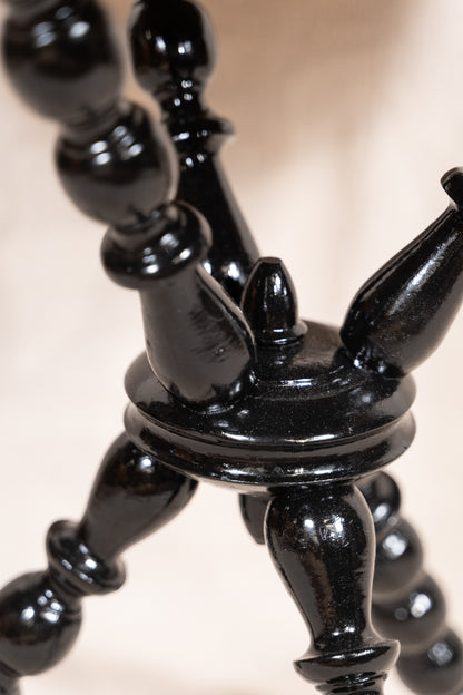 Close-up of a glossy black wooden object with turned, bulb-like legs and a circular base, photographed against a soft warm beige background.