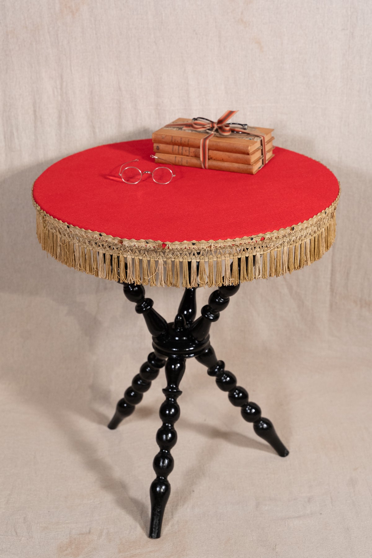 A round side table with a bright red upholstered top, beige fringe trim around the edge, and three black turned legs. A wrapped bundle of books and a pair of round glasses sit on the tabletop against a neutral fabric backdrop.