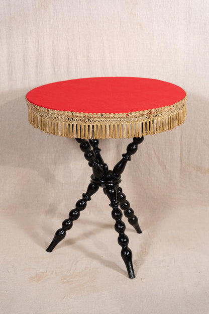 A round side table with a vibrant red upholstered top, a gold fringe trim, and a glossy black turned spindle legs base, set against a neutral beige fabric backdrop.