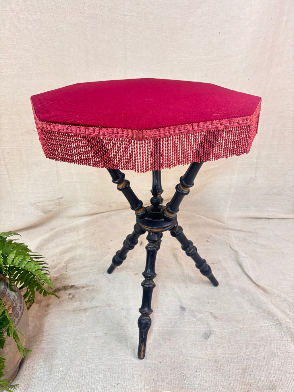 Antique Victorian ebonized bobbin-legged gypsy table with red velvet octagonal top and fringing, photographed against a cloth backdrop next to a plant