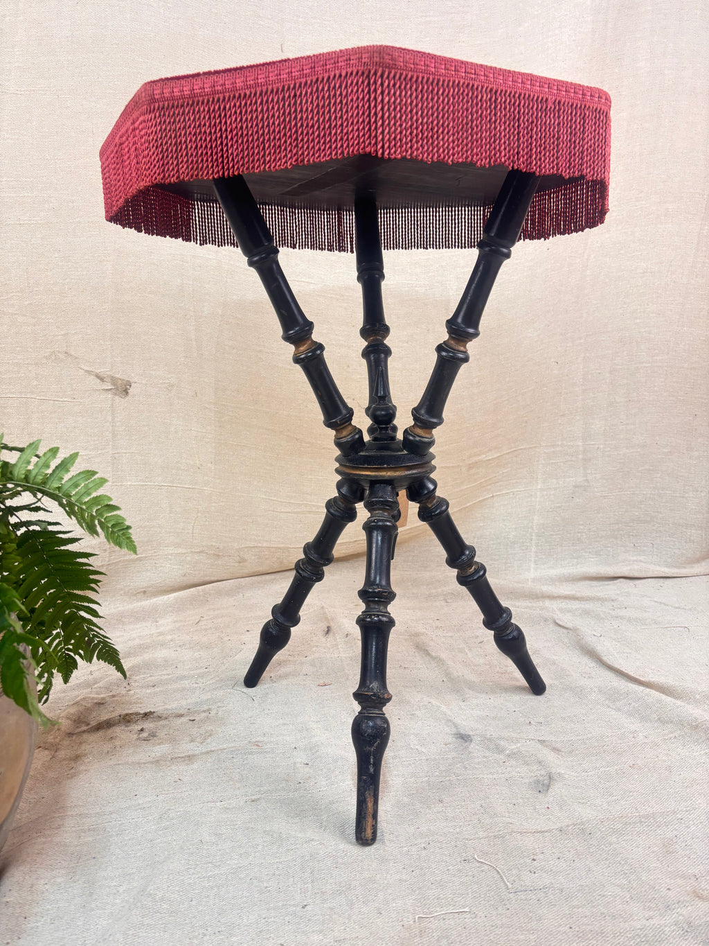 Antique Victorian ebonized bobbin-legged gypsy table with red velvet octagonal top and fringing, photographed against a cloth backdrop next to a plant