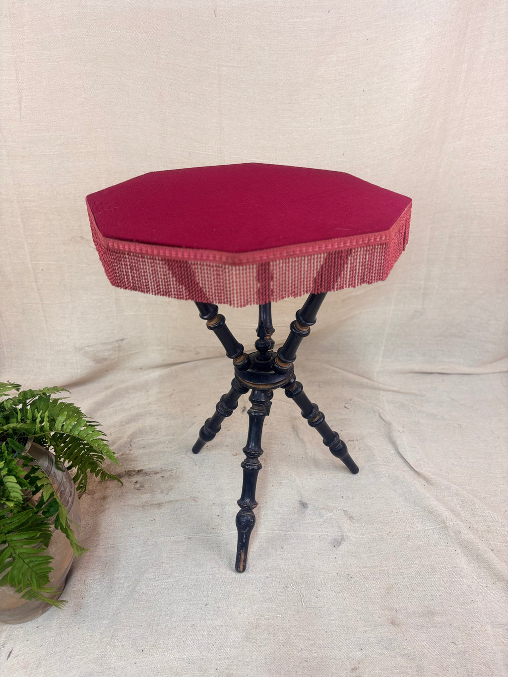 Antique Victorian ebonized bobbin-legged gypsy table with red velvet octagonal top and fringing, photographed against a cloth backdrop next to a plant