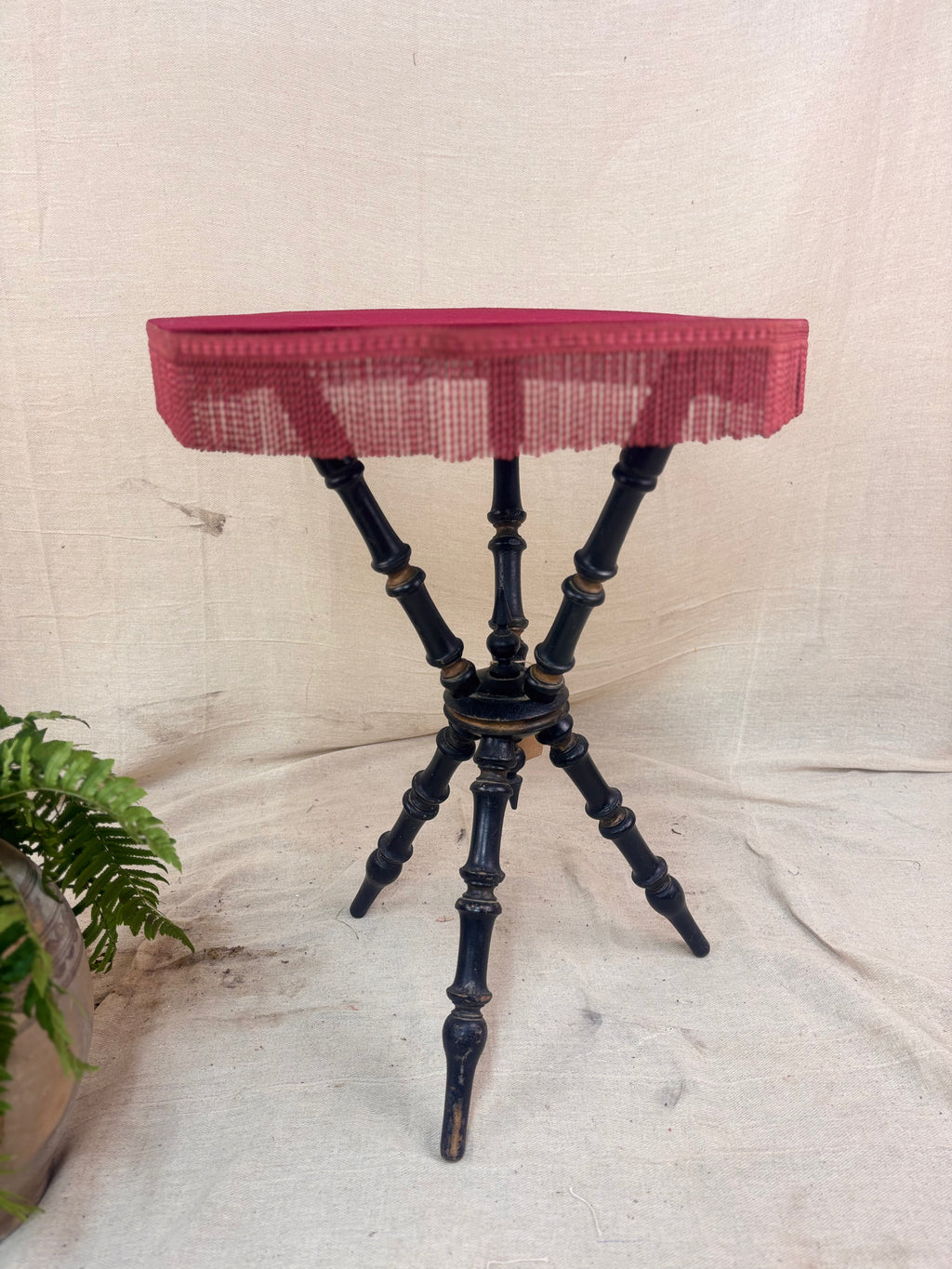 Antique Victorian ebonized bobbin-legged gypsy table with red velvet octagonal top and fringing, photographed against a cloth backdrop next to a plant