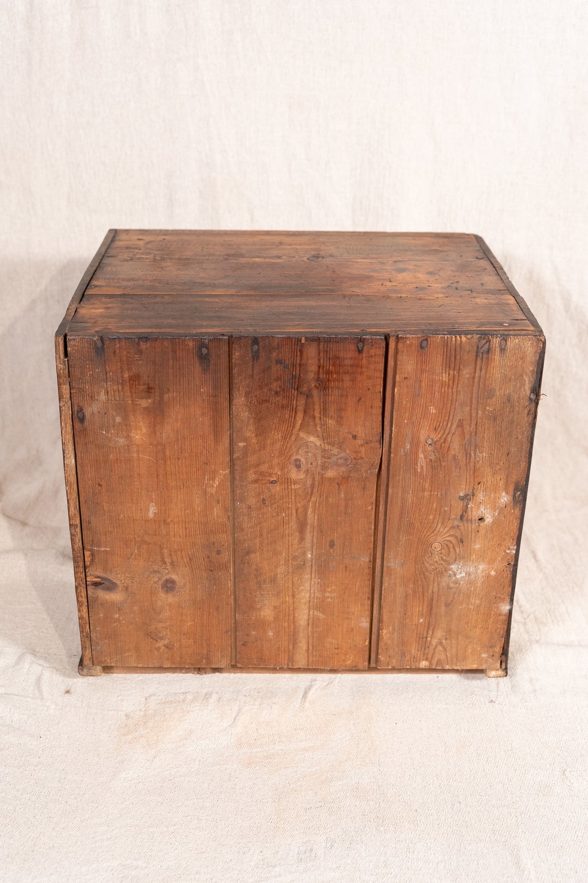 A rustic wooden storage chest or crate with three front doors, made from weathered brown wood planks, photographed against a neutral off-white fabric backdrop.