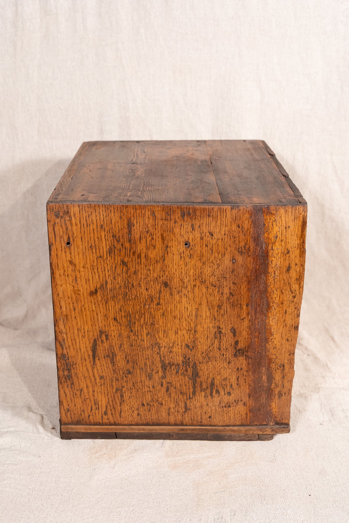 A vintage wooden storage chest or box made of weathered oak wood, showing a warm brown grain and dark distressing, photographed against a draped off-white fabric backdrop.