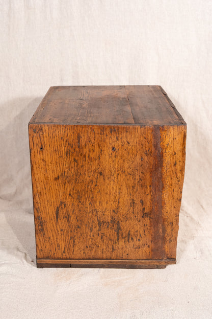 A vintage wooden storage chest or box made of weathered oak wood, showing a warm brown grain and dark distressing, photographed against a draped off-white fabric backdrop.