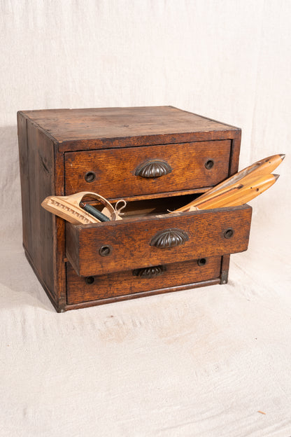 A vintage wooden chest of drawers with three drawers, one open, revealing wooden tools and a small scale inside. The chest has a warm brown wood finish with dark metal drawer pulls, set against a neutral off-white fabric backdrop.