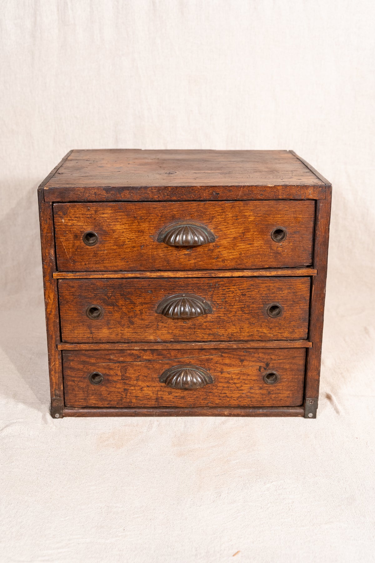 A small vintage wooden chest of three drawers with ornate metal handles, photographed against a neutral off-white fabric backdrop.