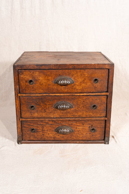 A small vintage wooden chest of three drawers with ornate metal handles, photographed against a neutral off-white fabric backdrop.