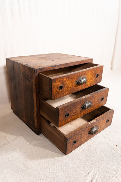 A rustic three-drawer wooden chest or organizer with weathered brown wood, dark metal round knobs, and open drawers showing light wood interiors, set against a draped off-white fabric backdrop.