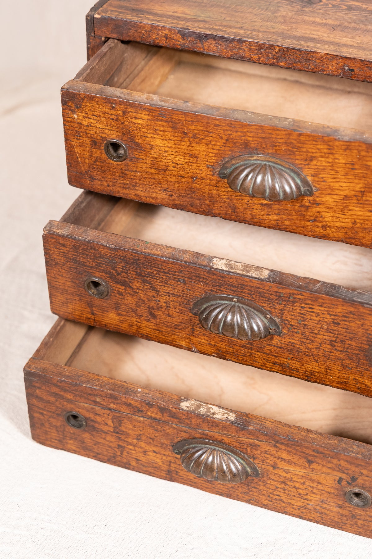 A close-up of a vintage wooden chest or cabinet with three stacked drawers pulled out, showing warm medium-brown wood grain and oval metal drawer pulls on a light neutral background.