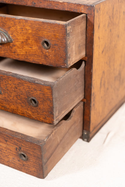 Close-up of a vintage wooden drawer chest with three partially pulled-out drawers, showing a warm brown wood grain and brass drawer pulls on a light fabric surface.