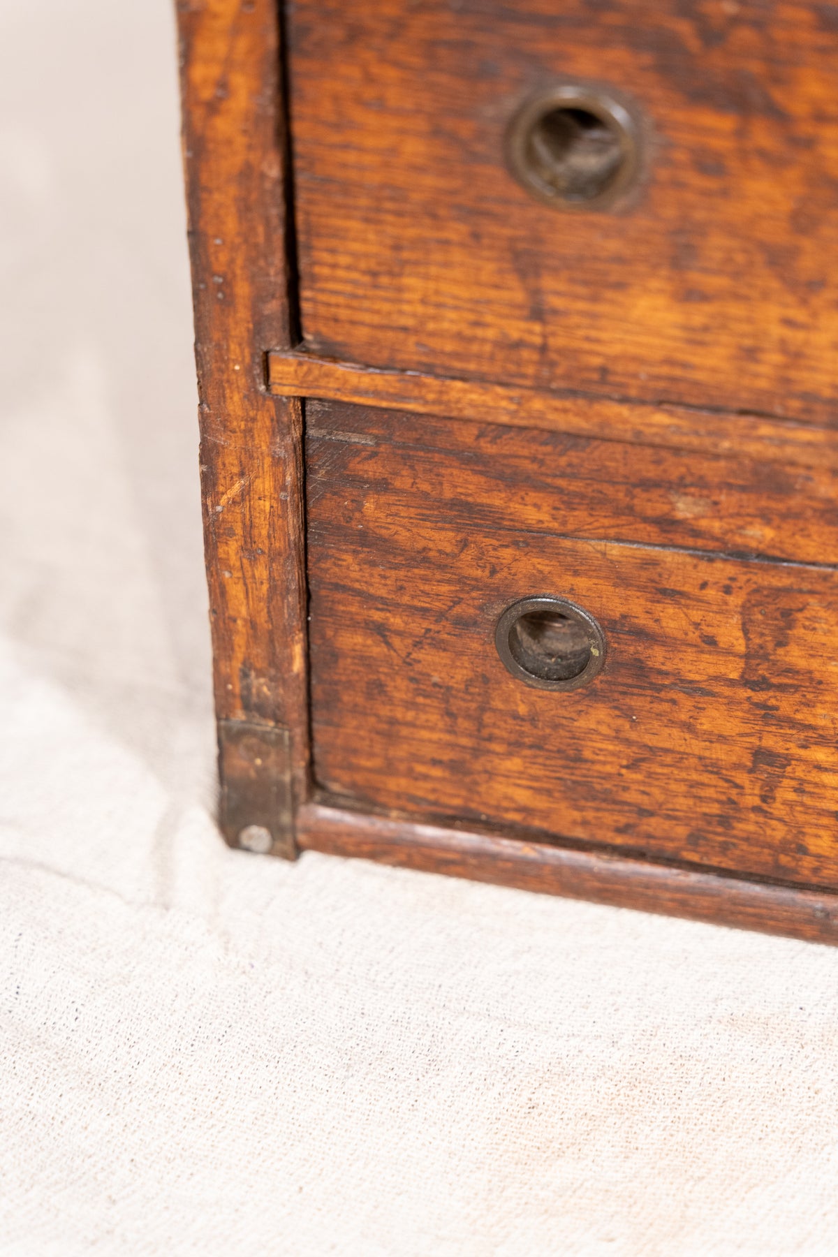 Close-up of a vintage wooden chest or cabinet with two visible drawers, showing the warm brown wood grain and round metal drawer pulls against a light fabric background.