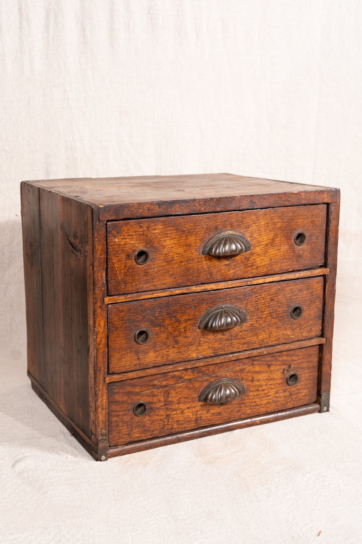 A rustic three-drawer wooden chest or side table made of dark brown oak, featuring vintage metal drawer pulls and brass corner brackets, set against a neutral off-white fabric backdrop.