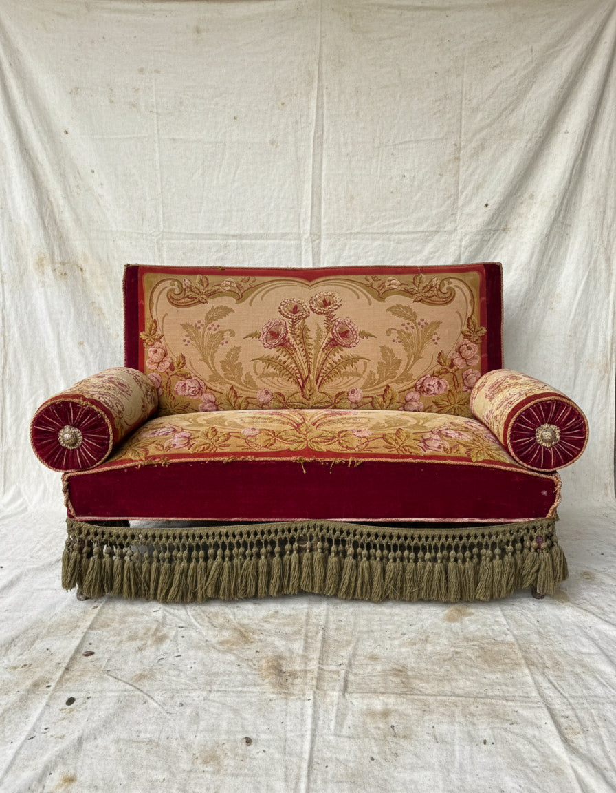 
An antique Victorian-style loveseat with red velvet trim, a beige floral tapestry back and seat, two red bolster pillows, and olive green fringe along the bottom.