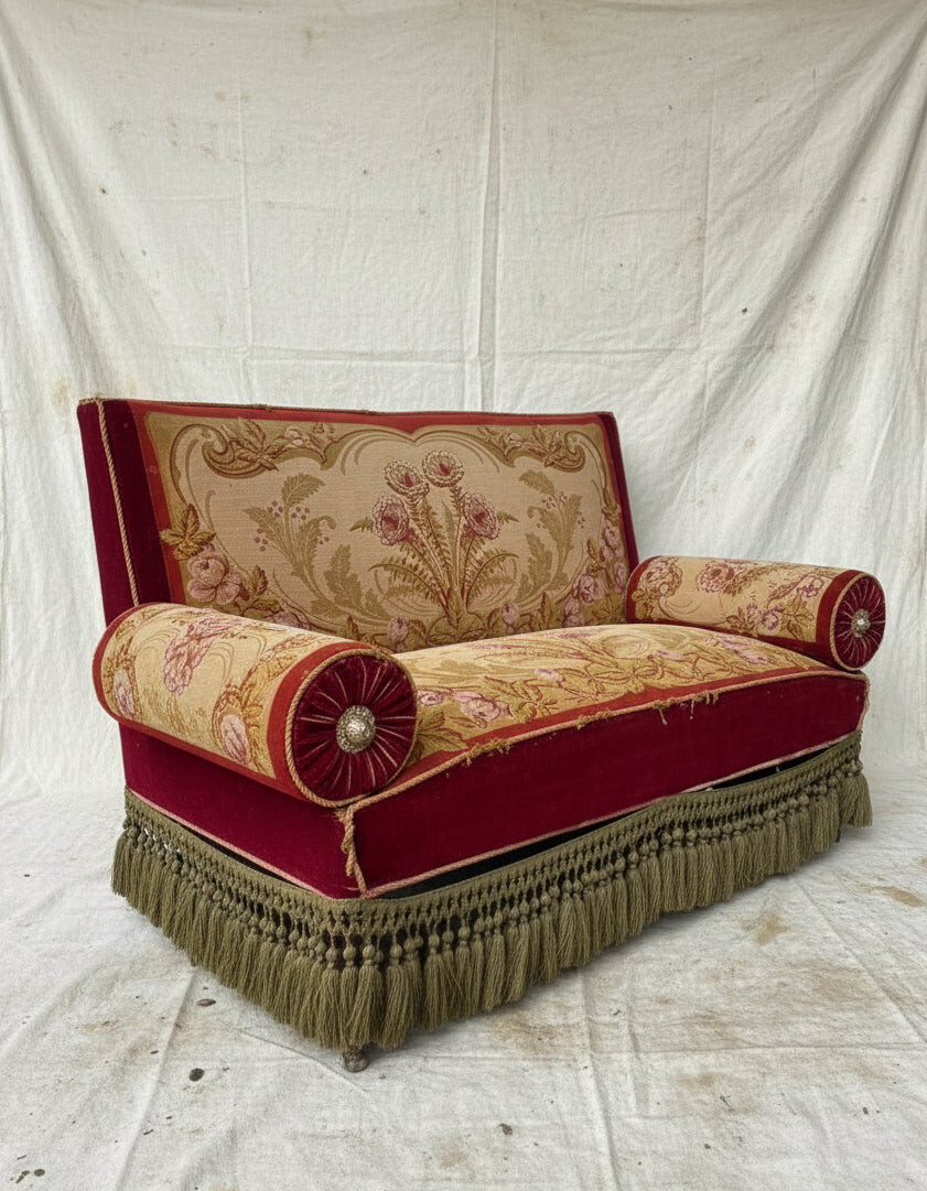 An antique Victorian-style loveseat with red velvet trim, a beige floral tapestry back and seat, two red bolster pillows, and olive green fringe along the bottom.